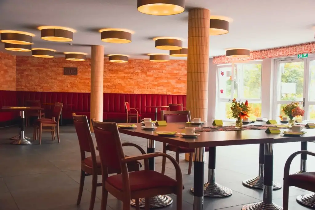 Cafeteria mit Tischen und Stühlen, runden Deckenleuchten und großen Fenstern mit Blick ins Grüne