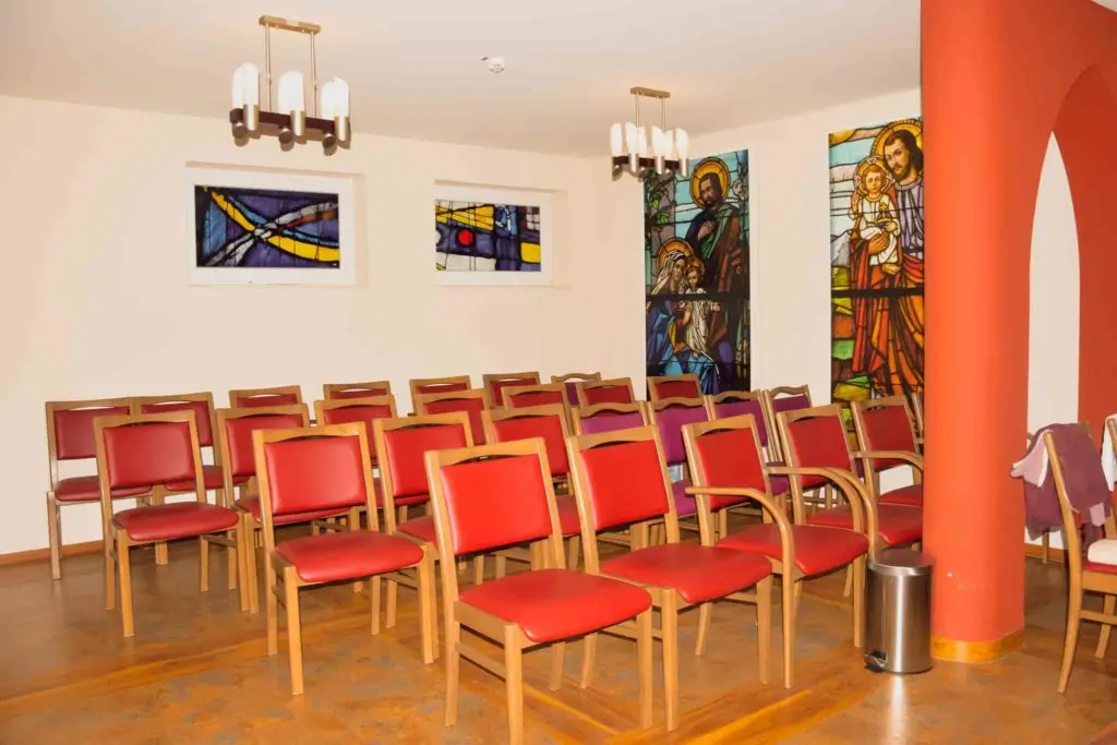 Gebetsraum mit mehreren roten Stühlen, bunten Glasfenstern mit religiösen Motiven