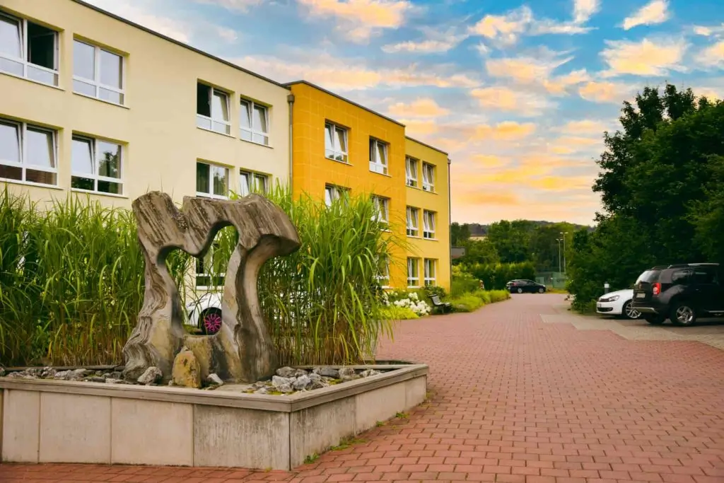 Skulptur aus Holz und Stein vor einem mehrstöckigen gelben Wohngebäude mit parkenden Autos auf gepflastertem Hof
