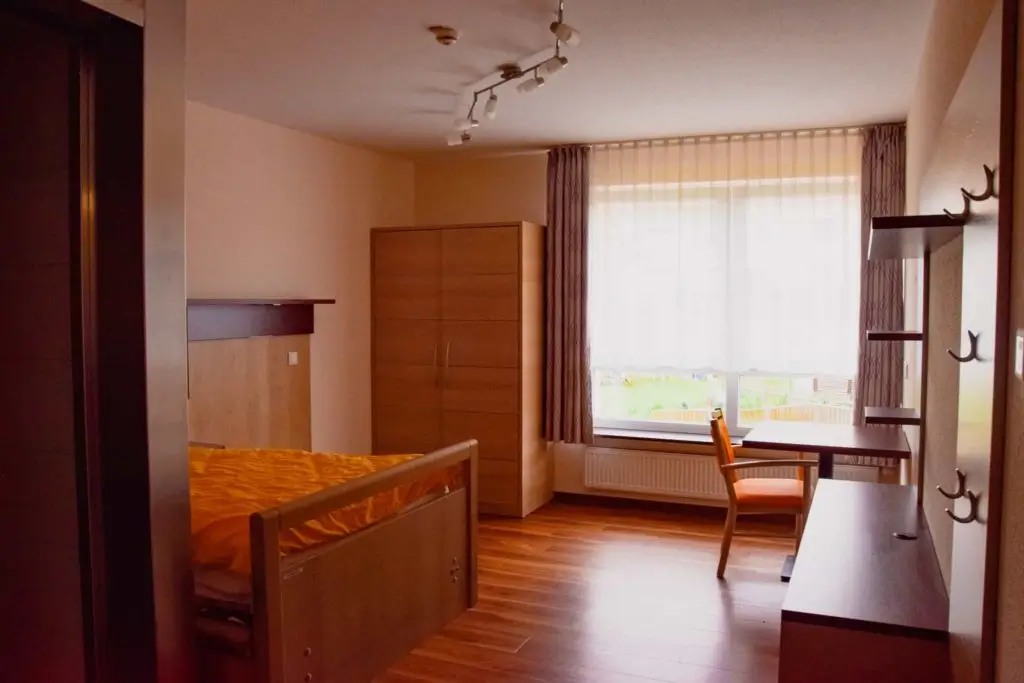 Ein schlicht eingerichtetes Zimmer mit einem Bett, einem Schrank, einem Schreibtisch mit Stuhl vor einem großen Fenster und Gardinen
