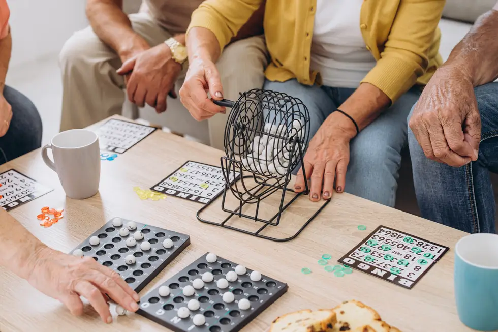 Mehrere ältere Personen sitzen an einem Tisch und spielen Bingo mit einem Metall-Bingo-Käfig und Spielkarten